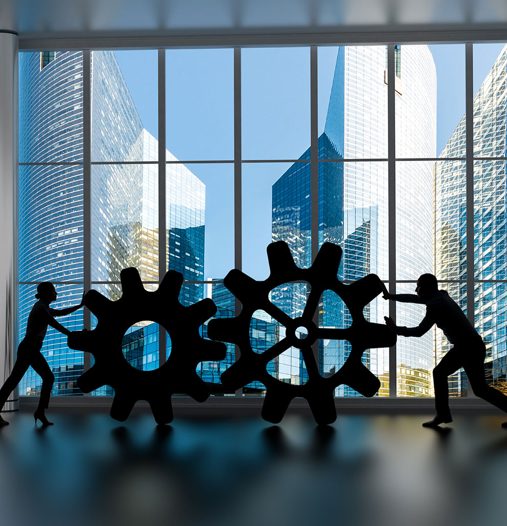 Shadows of woman and man pushing together giant gears in front of windows with sunlit corporate buildings in background